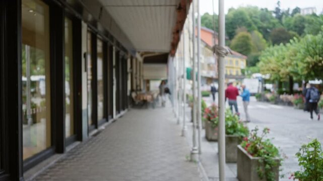 Covered arcade and shopfronts with soft defocused bokeh and blurry sidewalk leading past planters and trees, street; background backplate copyspace calm.