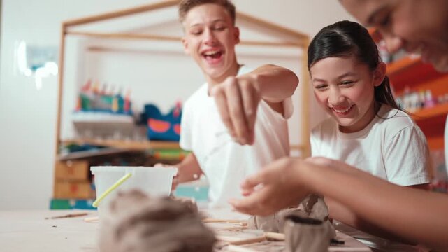 Handsome student dipping hand in to water to soften clay at art classroom. Group of diverse children working or modeling vase. Happy boy put water to young girl while laugh with happy. Edification