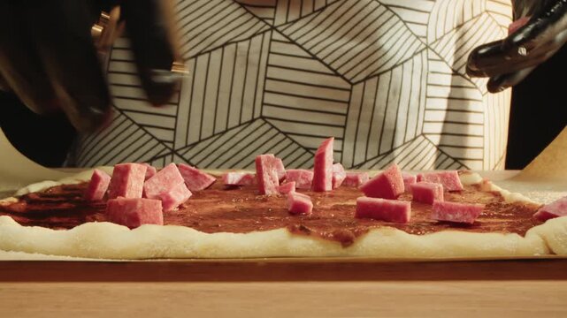 Closeup of pizza dough with tomato sauce and salami. Gloved hands of chef arranging sliced salami sticks on pizza base on baking paper. Man in apron stands behind wooden table in kitchen of restaurant