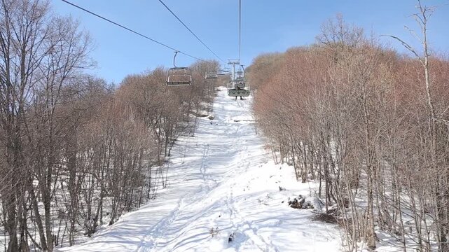 A scenic point of view shot from a ski chairlift descending a snow-covered mountain, passing bare trees, with majestic snow-capped peaks and a valley in the hazy distance.