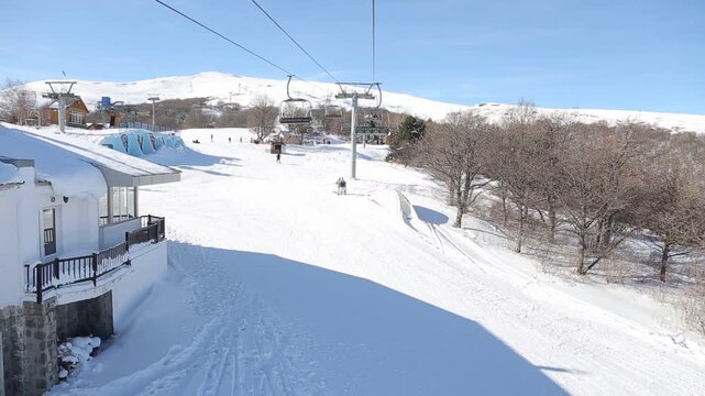 A scenic point of view shot from a ski chairlift descending a snow-covered mountain, passing bare trees, with majestic snow-capped peaks and a valley in the hazy distance.
