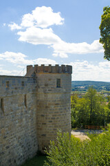 Sunlit stone tower rises above the old fortress wall in Altenburg, Germany. Soft clouds drift across a bright blue sky over rolling green hills beyond. Quiet paths and trees frame the historic scene. © serhii