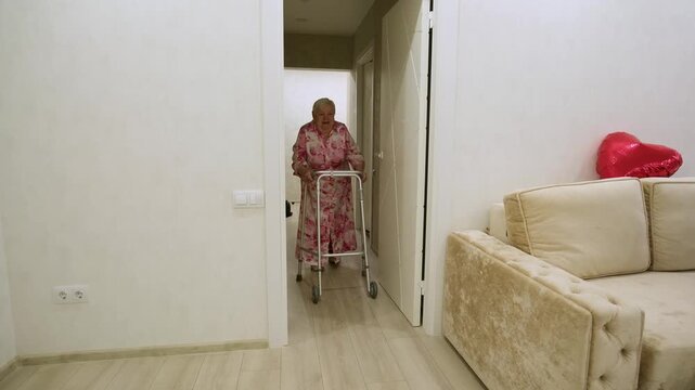 Senior woman with limited mobility using walking frame to move around her home, supporting herself while walking along a corridor towards the living room. Concept of aging and assisted living
