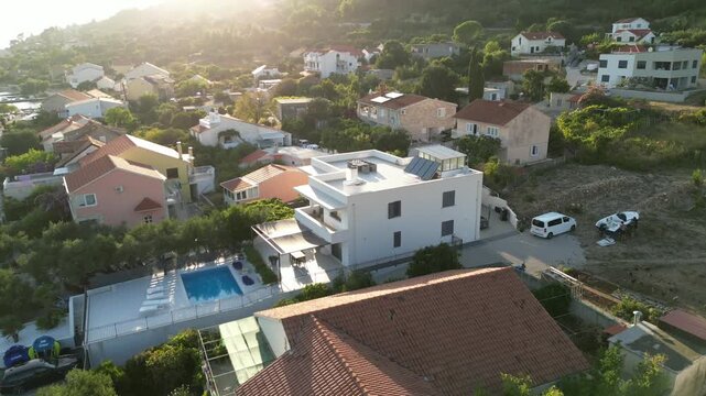 Aerial drone view of a modern white Mediterranean villa with a private pool at golden hour, surrounded by a hillside coastal neighborhood with lush greenery and terracotta roofs.