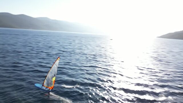 Aerial drone view of windsurfers gliding across deep blue Adriatic waters along the Croatian coast, with lush green hills and a clear sunny sky in the background.