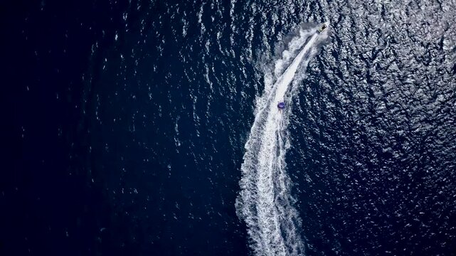 Cinematic top-down drone shot of a jet ski towing a rider across deep blue sea, carving a curved path with bright white wake and sparkling water texture.