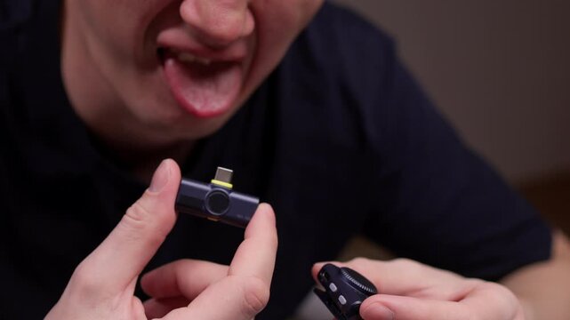 Man licking smartphone-compatible wireless microphone receiver while holding separate wireless lavalier microphone, demonstrating interaction with audio recording technology, close-up, slow motion.