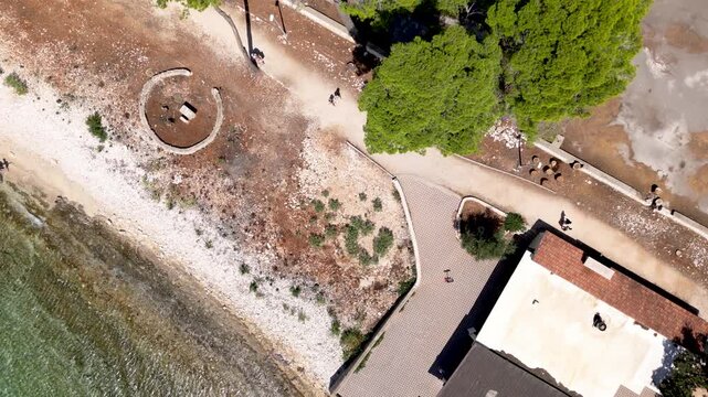Cinematic drone aerial over Badija Island, Croatia, gliding along a pine forest shoreline and pebble beach with a coastal path and dramatic mountains in the background.