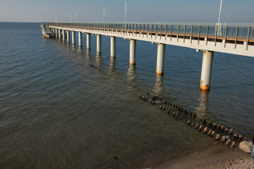 Fototapeta premium Long wooden pier extending over calm water with concrete supports and clear sky, ideal for fishing or leisure activities