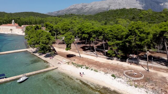 Cinematic drone aerial over Badija Island, Croatia, gliding along a pine forest shoreline and pebble beach with a coastal path and dramatic mountains in the background.