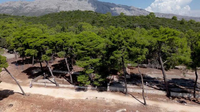 Cinematic drone aerial over Badija Island, Croatia, gliding along a pine forest shoreline and pebble beach with a coastal path and dramatic mountains in the background.
