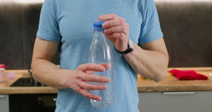 Young man twists small blue cap of empty plastic bottle. Male dressed in casual blue T-shirt stands in kitchen and unscrews plastic lid of transparent bottle