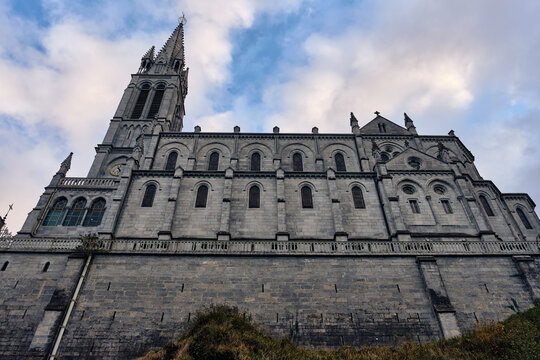 Lourdes, France - 19 November 2025: View of the imposing, grey Basilica of the Immaculate Conception, its towering spire piercing the cloudy sky from the elevated ground.