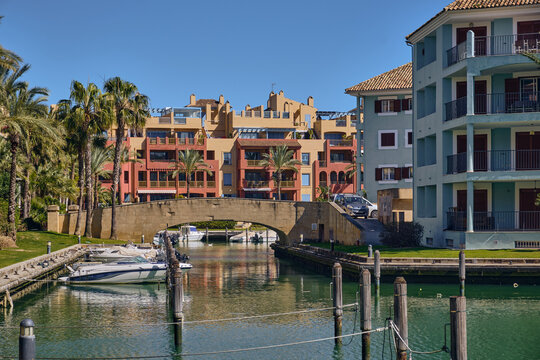Sotogrande, Spain - 22 February 2026: View of a tranquil canal reflecting the warm hues of terracotta buildings and the cool blues of the sky, framed by lush palms and a charming stone bridge.