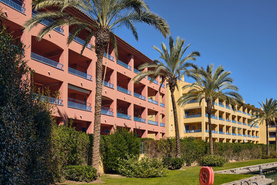 Sotogrande, Spain - 22 February 2026: View of peach and yellow buildings rise behind lush green foliage, punctuated by the slender silhouettes of palm trees against a clear blue sky.