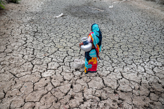 Satkhira, Bangladesh - 25 April 2025: View of a lone woman carrying water vessels across a parched, cracked earth, her vibrant clothing a stark contrast to the arid landscape.