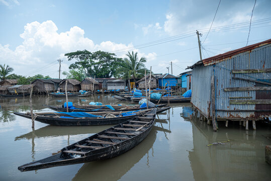 Khulna, Bangladesh - 12 July 2025: View of wooden boats floating on calm waters, reflecting the stilt houses and corrugated metal structures along the riverbank.
