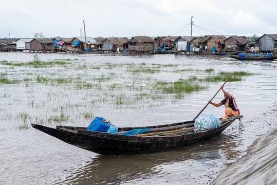 Khulna, Bangladesh - 13 July 2025: View of a weathered wooden boat pulled to the muddy bank, a woman with an oar facing stilt houses under a cloudy sky.