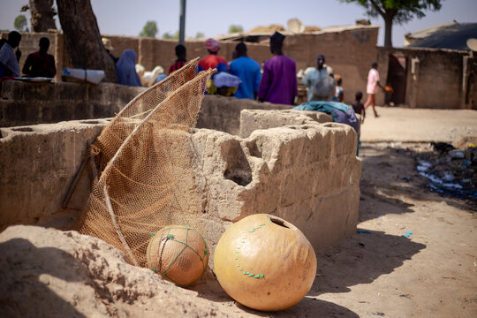 Argungu, Nigeria - 12 February 2026: View of sun-drenched earthen structures, with fishing nets and gourds resting in the foreground, against a backdrop of villagers and traditional buildings.
