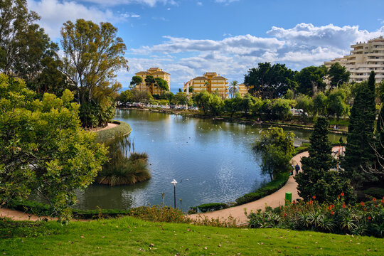 Benalmadena, Spain - 24 December 2025: View of Parque de la Paloma's tranquil lake reflecting the clear sky, embraced by vibrant green foliage and distant buildings.
