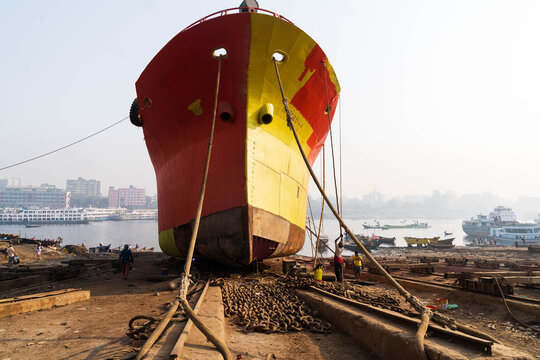 Keraniganj, Bangladesh - 11 February 2023: View of a massive, red and yellow ship's bow dominates the foreground, contrasting with the distant city skyline and the muddy, textured shipyard ground.