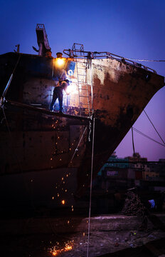 Keraniganj, Bangladesh - 13 December 2023: View of a shipbuilder amidst sparks against the stark silhouette of a vessel under the twilight sky, a scene of industry and labor.