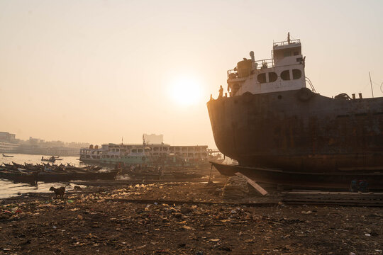 Keraniganj, Bangladesh - 11 February 2023: View of hulking ship beside smaller boats along the shoreline, reflecting the golden sunlight in a poignant scene.