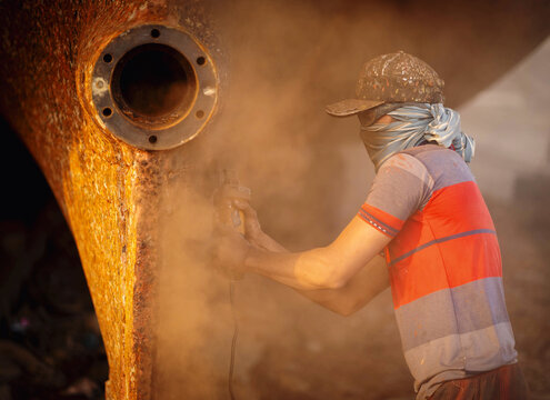 Keraniganj, Bangladesh - 11 February 2023: View of a worker grinding metal on a large rusty ship part, creating a cloud of dust in the shipyards of Keraniganj.