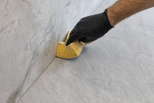 Worker in a black nitrile glove cleaning grout residue from gray porcelain tiles with a yellow sponge during kitchen renovation. Construction finishing and interior renovation