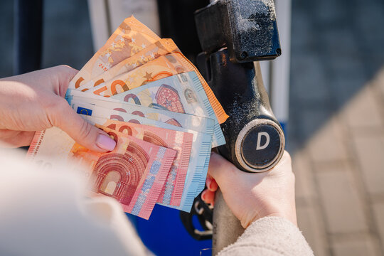 Hand holding various Euro banknotes near a fuel pump nozzle at a gas station, showcasing currency exchange for refueling vehicle