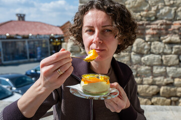 Fototapeta premium a woman with curly hair wearing a brown jacket, looking toward the camera as she holds a spoonful of layered passion fruit dessert from a glass jar, 