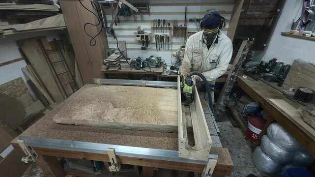 Frontal Static Portrait of Professional Joiner in Coverall and Respirator Surfacing Wood Slab with Router and Active Dust Extraction Slow Motion