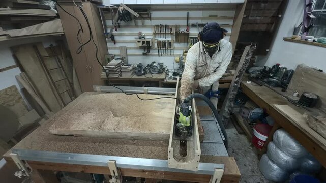Frontal Static Shot of Focused Carpenter in White Protective Suit Surfacing Wood Slab with Router Real Time Authentic Organized Workshop Pegboard Background