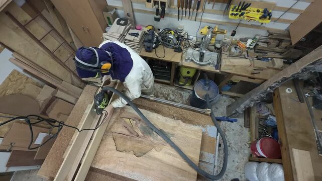 High Angle Panning Shot of Carpenter in Extreme Safety Hood Surfacing Wood Slab with Router Cluttered Workshop Slow Motion