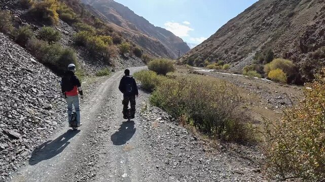 Two men riding electric unicycles along a gravel mountain road with rocks in Kyrgyzstan. The scene shows rugged terrain, natural surroundings, and off-road adventure.