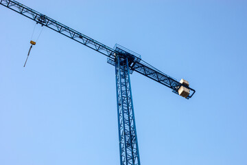 Naklejka premium construction tower crane with long horizontal jib and hook suspended in air, industrial machinery on building site under clear blue sky