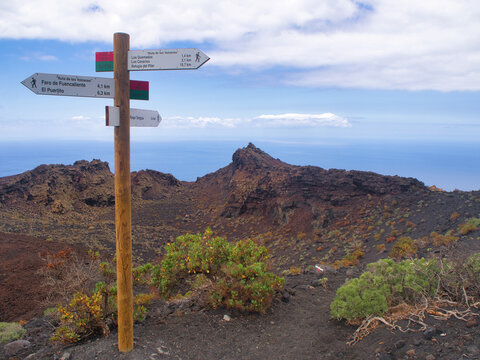 Hiking sign on the Volcano Route, La Palma - A wooden trail marker with directions and distances for the Ruta de los Volcanes hike, set against volcanic black earth and the ocean.