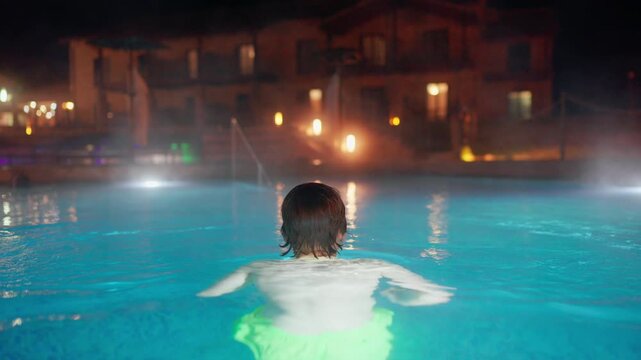 A person seen from behind relaxes in a steaming outdoor thermal pool at night. The illuminated turquoise water glows against the backdrop of a warmly lit luxury hotel or spa resort. Peaceful scene.