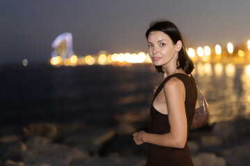 Fototapeta premium Female tourist is exploring citys attractions, brightly lit by electricity. Woman at waters edge, hotel skyscraper Arts in background. Night fell on city..