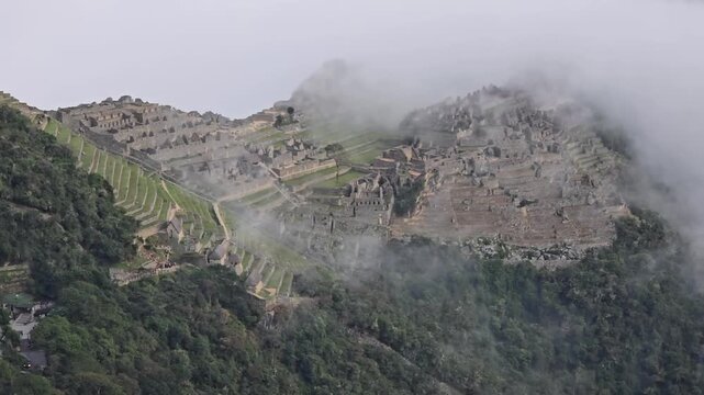 Machu Picchu, Peru - 26 June 2024. Slow pan across ancient Inca ruins and terraced slopes as low clouds move between stone structures on a steep mountainside.
