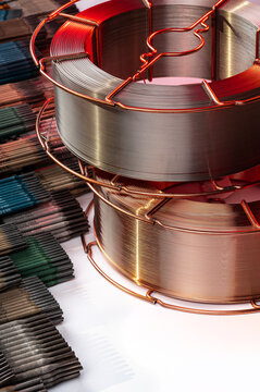 Vertical shot of welding wire spools and electrodes stack. Stacked reels of welding wire next to rows of color coded welding electrodes on white.