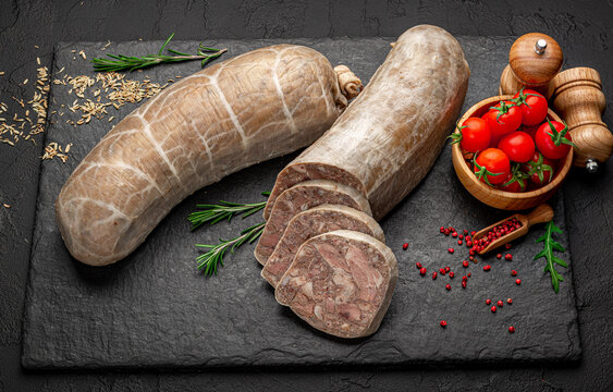 Sliced homemade headcheese meat terrine with spices on black board. Traditional meat jelly roll with rosemary, tomatoes and peppercorns on dark stone background