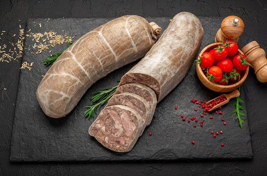 Homemade sliced meat jelly roll with spices on dark board. Cold meat appetizer with rosemary, tomatoes and pink pepper on black slate