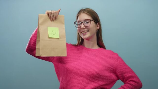 Woman holding a brown paper bag with green sticky note and smiley face in blue studio while smiling at camera; happiness.
