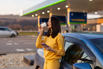 Fototapeta premium Coffee before miles. Woman driver enjoying morning coffee, standing outside her car in front of gas station, using cellphone to check the route