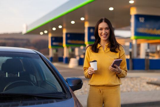 Young European woman with coffee cup and cellphone standing near her car against gas station, smiling at camera. Highway message pause concept