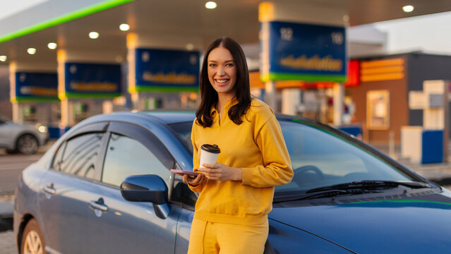 Happy traveler woman enjoying quick coffee break beside her car at modern gas station, smiling at camera, panorama shot