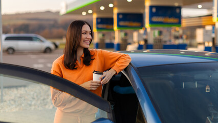 Naklejka premium Happy woman standing by her car at gas station, holding coffee cup in hands, looking aside and smiling, taking break during road trip, panorama shot