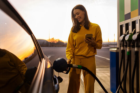 Happy woman driver filling her car with fuel at the gas station and using cellphone, paying online using app on phone