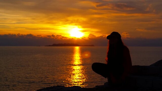 Young woman silhouette watching a golden sunset over the ocean. Silhouette of a pensive young woman with a baseball cap sitting on a rocky shore, contemplating the breathtaking golden sunset